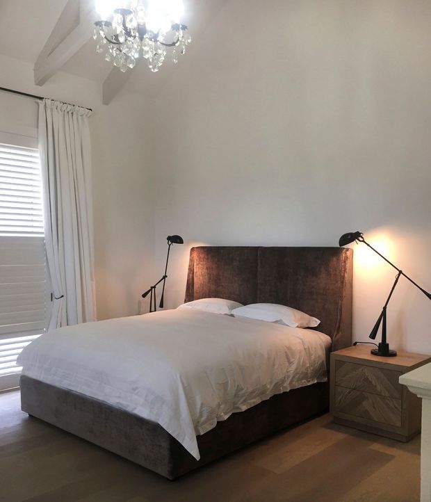 A refined bedroom with a large upholstered bed, white bedding, matching bedside tables and adjustable black reading lamps. The room features pale walls, exposed painted ceiling beams, timber flooring, plantation shutters, full-length curtains and a crystal chandelier, creating a soft, elegant atmosphere.