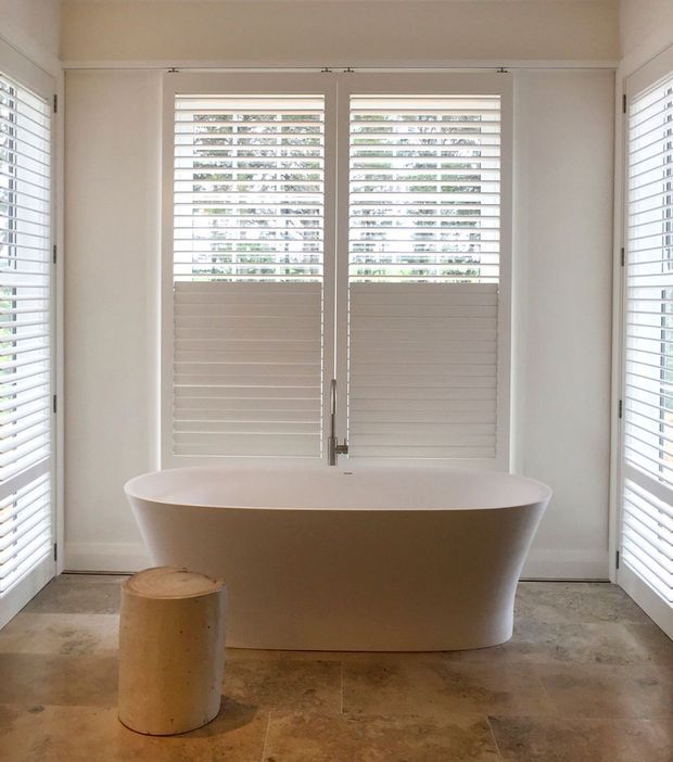 Bright, refined bathroom featuring a freestanding white bathtub positioned in front of shuttered windows, with plantation shutters on three sides, pale walls and trim, and a warm beige stone-tile floor. A small cylindrical timber stool sits beside the bath.