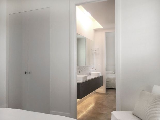 A bright minimalist bedroom viewed toward an open bathroom doorway. The bedroom has white walls, a tall built-in white wardrobe, and the edge of a white bed and upholstered seating. The adjoining bathroom features a dark floating vanity with twin white vessel basins, mirrored cabinetry, pale wall finishes, and warm stone-look flooring. A partially visible bed appears beyond the bathroom area.