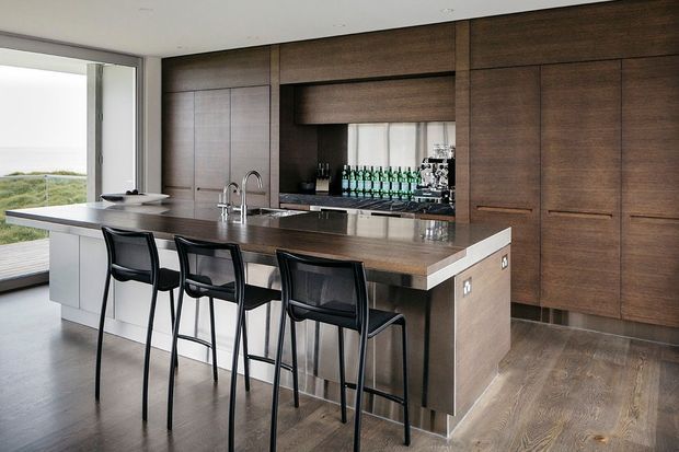 Contemporary kitchen with a large island and four black bar stools, dark timber wall-to-wall cabinetry, a reflective stone benchtop, integrated sink and tapware, and a mirrored or glazed drinks display niche. Floor-to-ceiling glazing opens to a coastal landscape beyond.