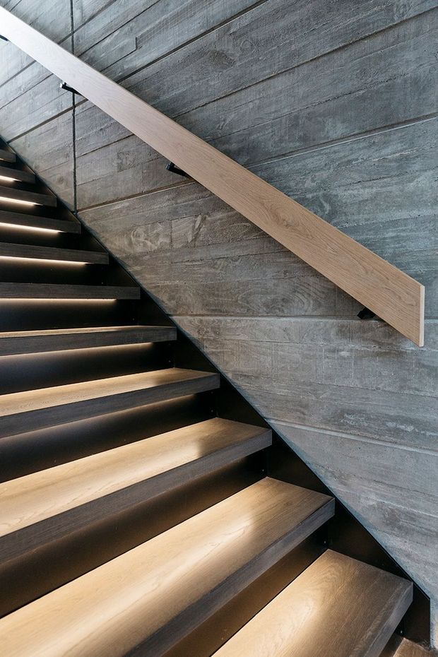 Close-up interior view of a contemporary stairway with broad dark timber treads, black structural risers and integrated warm LED lighting. A pale timber handrail contrasts against a textured grey concrete wall, creating a refined industrial-modern architectural detail.