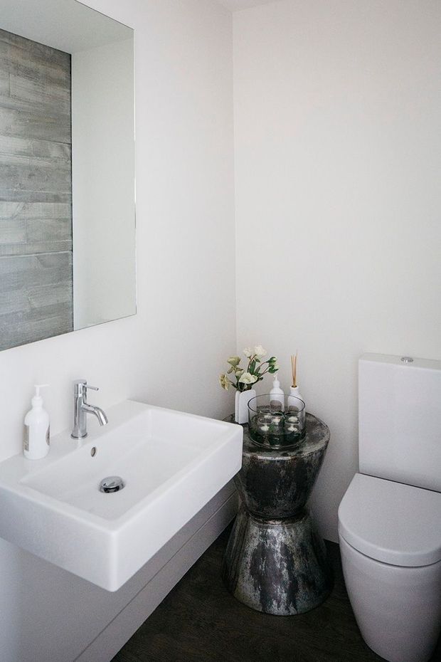 A compact contemporary bathroom featuring a wall-mounted white rectangular basin with chrome tapware, a white toilet, a large frameless mirror, white walls, dark wood-look flooring, and a dark metallic decorative pedestal beside the toilet.