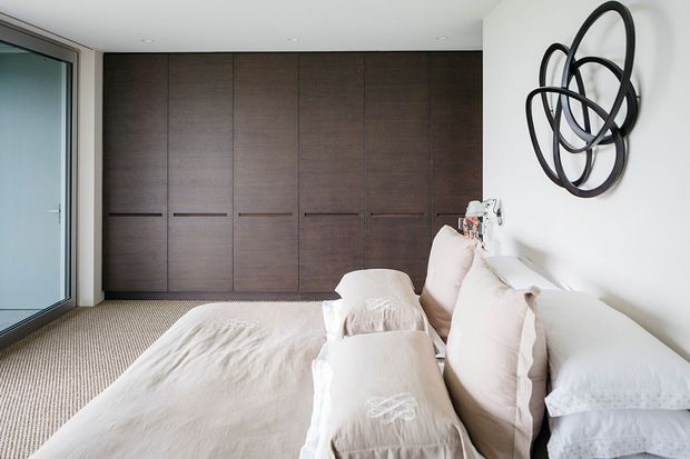 A bright, contemporary bedroom with a low-profile bed dressed in layered white and cream linen bedding. A full wall of dark timber built-in wardrobes provides strong contrast against the white walls, while a large abstract black sculptural artwork hangs above the bed. A glazed door or window is visible at left, and the room has soft neutral carpeting.