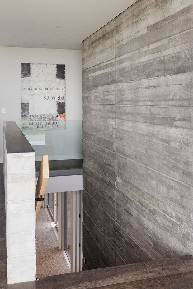 Interior view of a contemporary stairway or landing with a dark timber stair or floor surface, a large textured grey concrete-look feature wall, white painted walls, glass balustrade, and a partial view down to glazed doors and lower-level circulation. An abstract artwork is visible on the far wall.