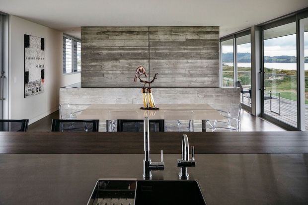 Open-plan contemporary kitchen and dining area with a dark island benchtop and undermount sink in the foreground, a long dining table with transparent chairs, and a textured horizontal timber feature wall behind a raised bench. Full-height corner glazing opens to a green waterside landscape and provides abundant natural light.