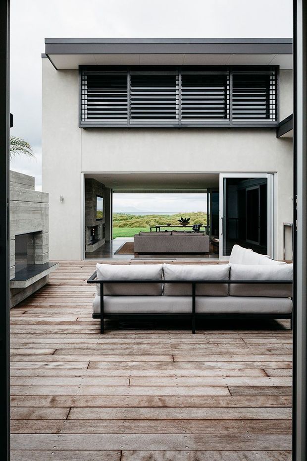 A contemporary coastal-facing home viewed from a timber deck, with an outdoor lounge sofa in the foreground, a built-in outdoor fireplace at left, and large sliding openings connecting to an interior living room. The facade is rendered in light tones with dark aluminium-framed windows and horizontal louvres beneath a projecting flat roof.