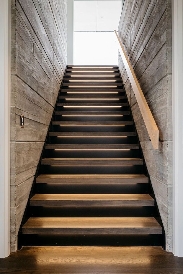A narrow, straight stairway with open-riser timber treads set into a dark metal stair structure. The stairwell is enclosed by textured horizontal timber-clad walls, with a simple timber handrail on one side and bright natural light at the upper landing. The design has a restrained, contemporary material palette of dark metal, warm wood, and concrete-like grey surfaces.