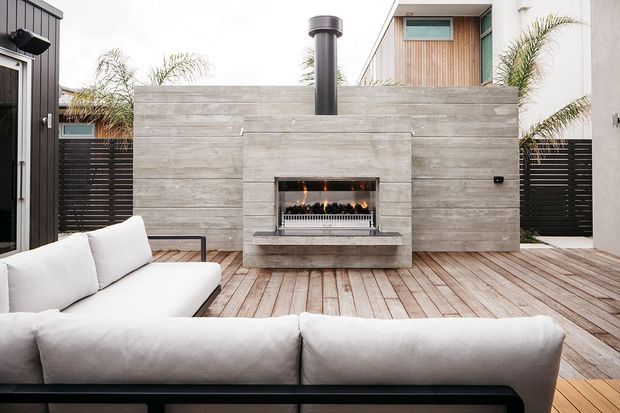 A contemporary outdoor entertaining area with a timber deck, built-in gas fireplace clad in horizontal grey concrete-look panels, black flue, and white outdoor sectional seating. Dark horizontal privacy fencing, timber-clad upper-level walls, and tropical planting surround the courtyard-like space.