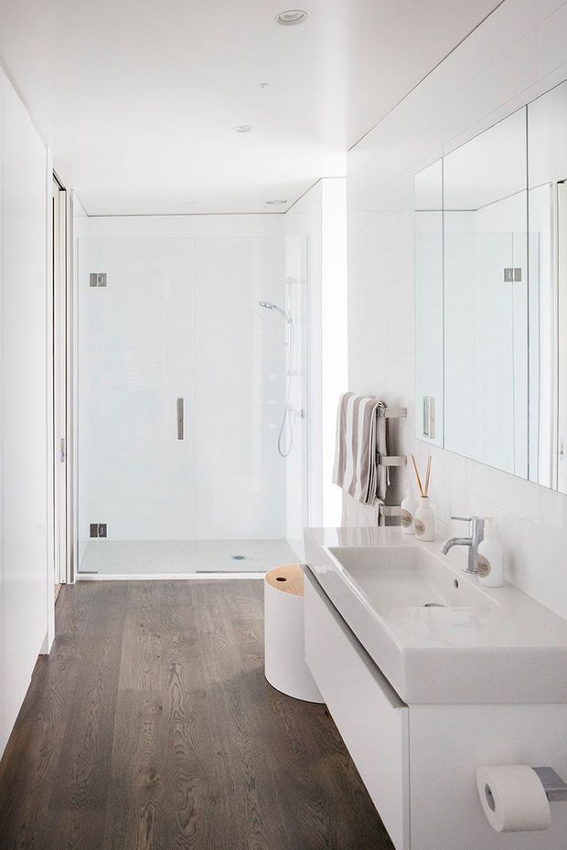 Bright contemporary bathroom with a large frameless glass walk-in shower, double vanity with integrated basins, mirrored wall cabinets, white tiled walls, recessed ceiling lights, and dark timber-look flooring. Neutral towels and minimal accessories add subtle warmth.