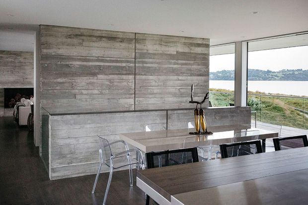 Modern open-plan dining area featuring a long dark timber dining table, transparent acrylic dining chairs, and a large built-in grey concrete or cement-look wall and bench with integrated fireplace niche. Floor-to-ceiling glazing overlooks a waterfront landscape, while a living area is partially visible through the opening at left. The space has a restrained, minimalist material palette and strong horizontal lines.