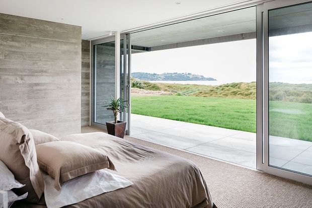 A serene contemporary bedroom with a low-profile bed dressed in neutral taupe bedding, a textured concrete-look feature wall, and expansive floor-to-ceiling sliding glass doors opening onto a lawn and coastal landscape. The room has a soft, natural palette and strong indoor-outdoor connection.