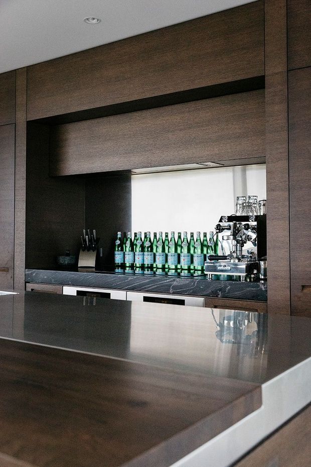 A sophisticated built-in home bar or hospitality counter clad in dark timber joinery, with a dark stone benchtop, under-cabinet shelving, a commercial espresso machine, glassware, and rows of bottled water. A broad reflective counter occupies the foreground, creating a refined, contemporary bar setting.