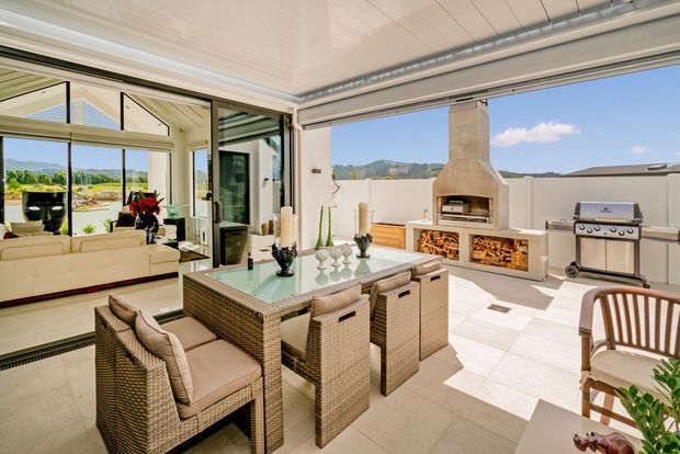Bright indoor-outdoor entertaining area with a large dining table and woven dining chairs, opening through wide glazed doors to a living room and scenic landscape. The tiled terrace includes a built-in masonry fireplace with integrated wood storage and adjacent stainless-steel barbecue, creating a refined outdoor dining and cooking space.