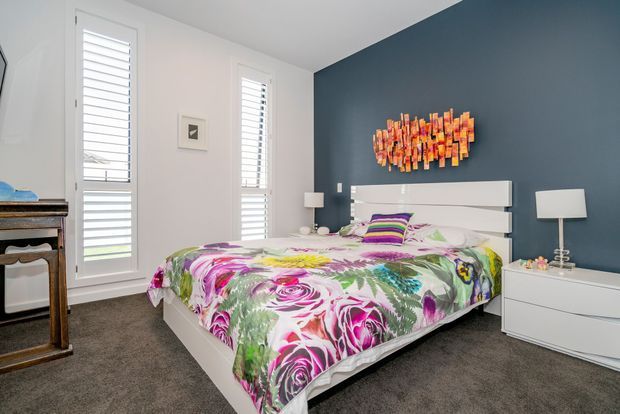 Bright contemporary bedroom with a white platform bed, colourful floral bedding, dark blue feature wall, white bedside tables and lamps, plantation shutters, and grey carpet. A vibrant abstract wall artwork adds a strong accent above the bed.