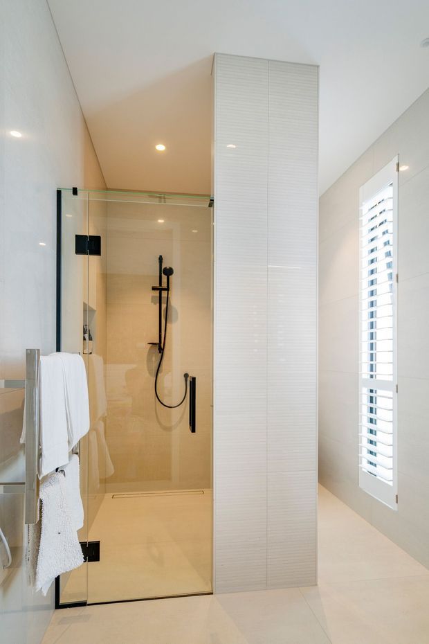 Bright, contemporary bathroom featuring a frameless glass walk-in shower with matte black fixtures, pale tiled walls and floor, a tall white ribbed privacy/storage panel, and a white shuttered window. A towel rail with white towels is visible beside the shower.