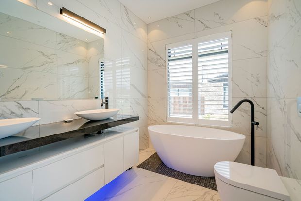 Bright contemporary bathroom with full-height white marble-look wall and floor tiling, a floating white vanity with dark stone benchtop, vessel basin, freestanding oval bathtub, matte black tapware, wall-mounted toilet, mirrored cabinetry, and a shuttered window. Blue LED lighting accents the underside of the vanity.