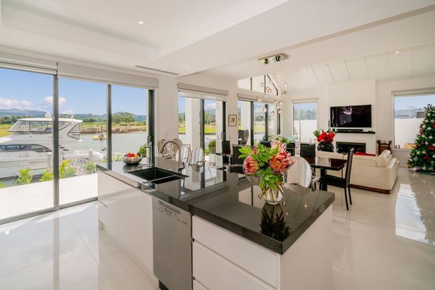 Bright open-plan kitchen and living area with a large central island, glossy dark benchtops, white cabinetry, extensive floor-to-ceiling glazing, and views toward a marina or waterfront outside. The living zone includes a television, fireplace or media unit, light upholstered seating, and festive floral and Christmas-tree decor.