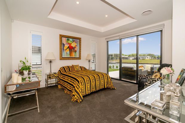 Bright, contemporary bedroom with a central bed dressed in a bold black-and-gold patterned bedspread, dark carpet, white walls, recessed tray ceiling, framed artwork, mirrored bedside tables, and large sliding glass doors opening to a balcony or terrace with views of a landscaped residential setting. A glass-topped console or vanity with decorative objects occupies the foreground.