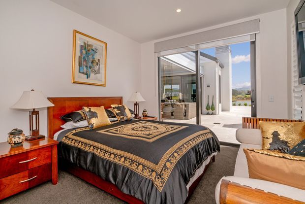 A bright, refined bedroom with a large bed dressed in a black and gold patterned coverlet, a rich reddish-brown timber headboard and matching bedside furniture, table lamps, framed artwork, and a light carpet. Full-height glazed doors open to a sunny outdoor terrace and reveal a contemporary adjoining space beyond.