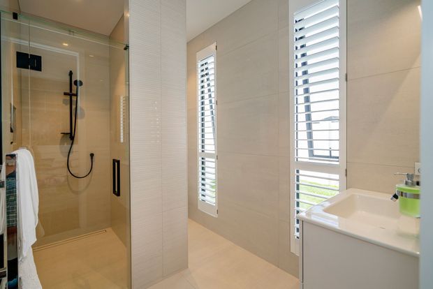Bright contemporary bathroom featuring a frameless glass walk-in shower with black fixtures, pale large-format wall and floor tiles, white louvered windows, and a white floating vanity with integrated basin. The space is clean, light-filled, and compact.