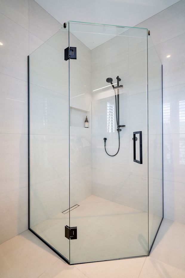 A bright, contemporary walk-in shower enclosure with clear frameless glass panels, black hardware, a black shower rail and handheld fixture, and large white wall and floor tiles. A recessed wall niche provides storage, while plantation shutters are visible through the glazing.