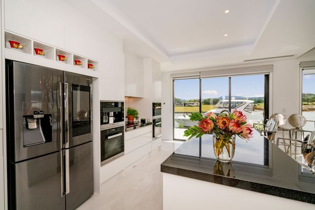Bright contemporary kitchen with extensive white built-in cabinetry, integrated stainless-steel appliances, a dark stone island benchtop, and large windows overlooking a landscaped suburban setting. A small dining area is visible at the right.