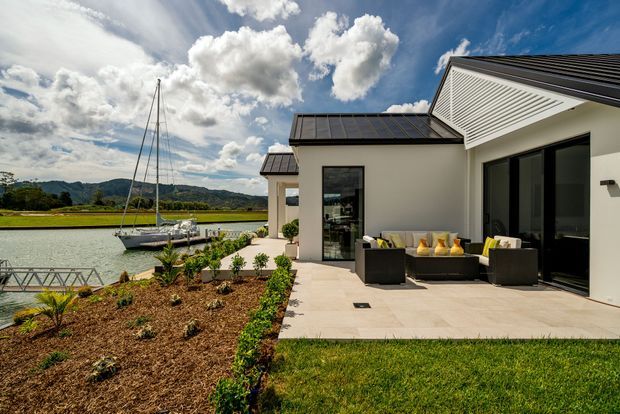 Contemporary single-storey waterfront home with a pale rendered exterior, dark pitched roof sections, expansive black-framed glazing and a tiled outdoor terrace. The terrace is furnished as an outdoor lounge overlooking a marina or river, with landscaped garden beds and lawn in the foreground.