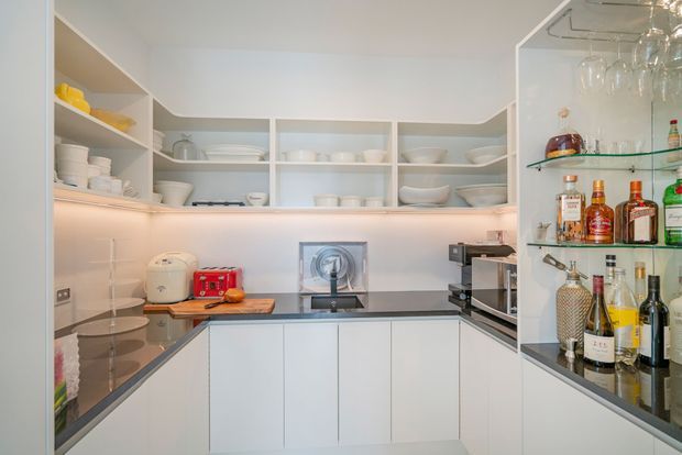 Bright contemporary galley-style kitchen with white handleless cabinetry, open shelving displaying crockery, dark stone-look benchtops, integrated appliances, and a glass-fronted drinks cabinet with glassware and bottles. Warm under-cabinet lighting adds a subtle glow.