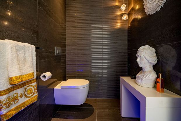 A dramatic, luxurious bathroom with dark glossy stone-look wall and floor finishes, a wall-hung white toilet, decorative patterned towels, warm pendant lighting, and a white sculptural bust displayed on a minimalist vanity or cabinet. A textured reflective feature wall adds depth and visual interest.