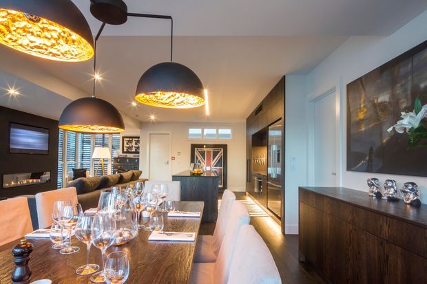 A sophisticated open-plan dining and living area with a long timber dining table, upholstered dining chairs, dark built-in cabinetry, a kitchen island, and a lounge zone with a fireplace and television. Dramatic black pendant lights with warm textured interiors illuminate the room, while artwork and decorative objects add refined character.