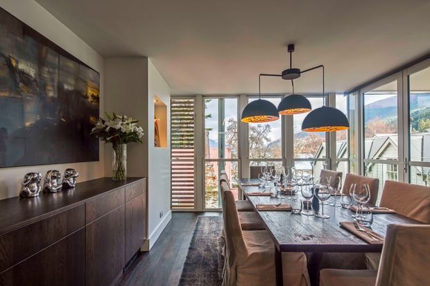 A refined dining room with a long set dining table, upholstered dining chairs, sculptural pendant lights, and floor-to-ceiling glazing overlooking a mountainous landscape. A dark timber sideboard, large abstract artwork, floral arrangement, and carefully set table create a warm, contemporary interior.