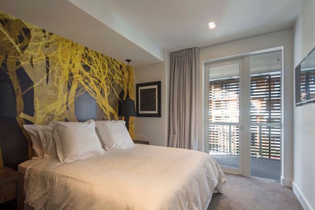 A contemporary bedroom with a large white upholstered bed, layered white bedding and pillows, and a dramatic black-and-gold botanical feature wall behind the headboard. Floor-to-ceiling curtains frame glazed balcony doors with horizontal timber blinds, while a framed artwork, bedside furniture, and recessed ceiling lighting complete the room.