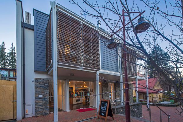 Exterior street-level view of a contemporary multi-storey commercial building with grey cladding, prominent timber louvre screens, stone-clad wall sections, large glazed openings, and a recessed illuminated ground-floor entrance. The building sits along a paved pedestrian frontage with street trees and signage.