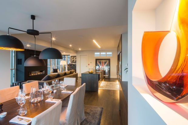 A refined open-plan dining and living area with a warm, contemporary interior. The foreground features a formal timber dining table set with glassware and upholstered dining chairs, while the background includes a seating area, dark suspended pendant lights, a central kitchen island or console, recessed and linear lighting, timber flooring, and a sculptural orange wall artwork.