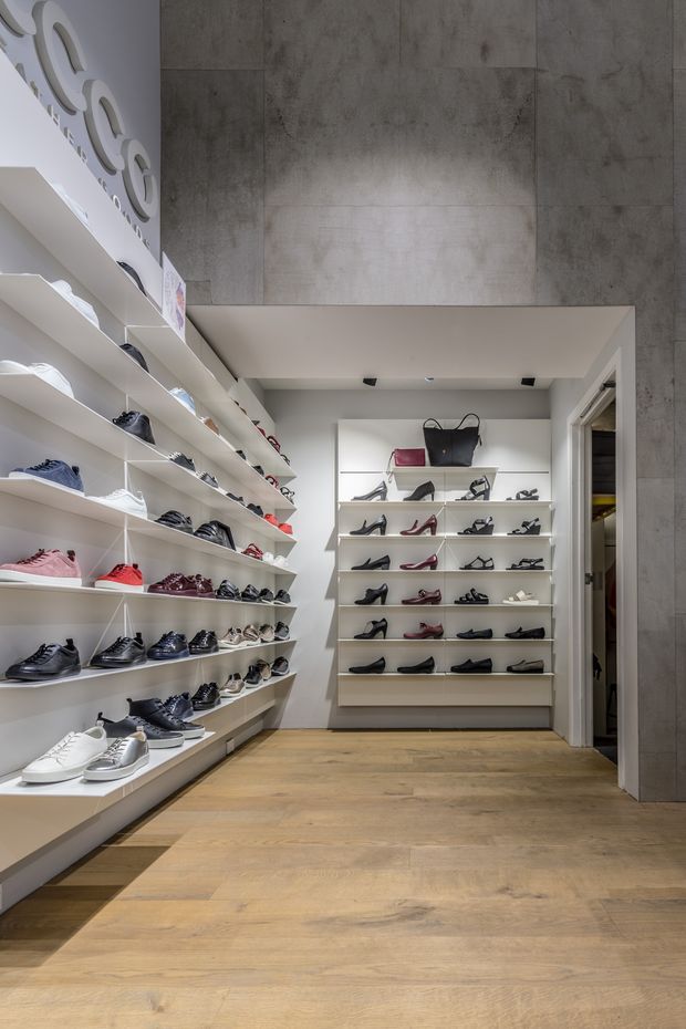 A modern retail shoe and accessories showroom with extensive white built-in display shelving along the walls, showcasing shoes and handbags. The space has a warm timber-look floor, large grey concrete-look wall tiles, white painted shelving and ceiling, recessed lighting, and a clean, minimalist commercial fit-out.