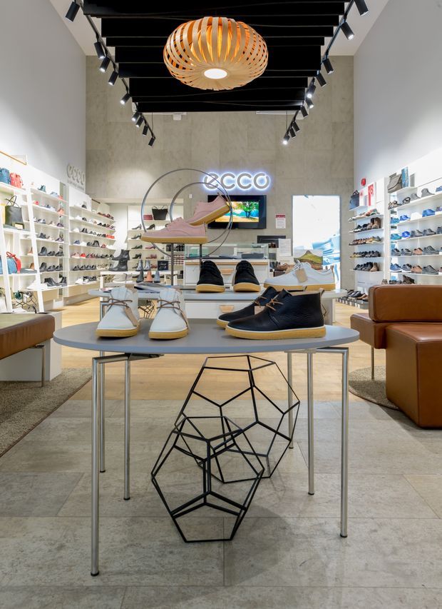 Interior view of a contemporary ECCO footwear retail store, with shoes displayed on white wall shelving and central tiered display tables. The space features a pale stone or concrete-look tiled floor, light textured walls, a black exposed-beam ceiling with track lighting, a sculptural wood pendant, leather seating, and branded signage.