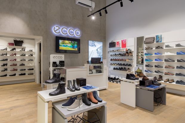A modern ECCO footwear retail store interior with extensive wall-mounted shoe displays, central white display tables, a checkout or service counter, timber-look flooring, and a concrete-textured feature wall with illuminated branding. Track lighting and wall shelving create a clean, bright commercial showroom environment.