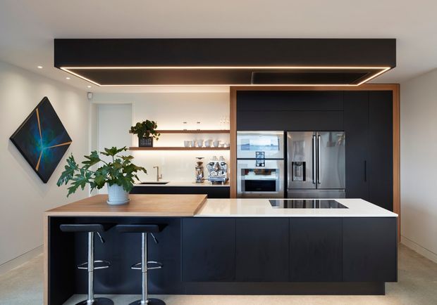 Modern kitchen with a large black island, white stone benchtop, integrated cooktop, timber breakfast-bar extension, black cabinetry, open timber shelving, stainless-steel appliances, and a dramatic recessed linear ceiling light. The space has a clean, contemporary fit-out with indoor plants and minimal furnishings.