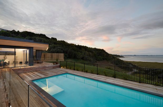A contemporary coastal residence viewed from an elevated outdoor deck at dusk. The image is dominated by a rectangular swimming pool with clear turquoise water, surrounded by timber decking and black metal balustrading. A glass-walled living area with warm interior lighting sits to the left, while dense coastal vegetation, shoreline, and calm water extend into the background.