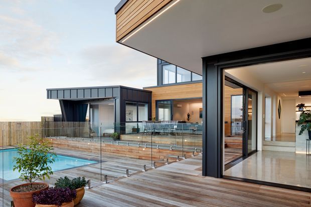 Contemporary multi-level home viewed from an outdoor deck, featuring a swimming pool enclosed by frameless glass balustrades, timber decking and cladding, extensive floor-to-ceiling glazing, and strong black-framed openings. The image looks toward an open-plan interior and covered outdoor entertaining area.
