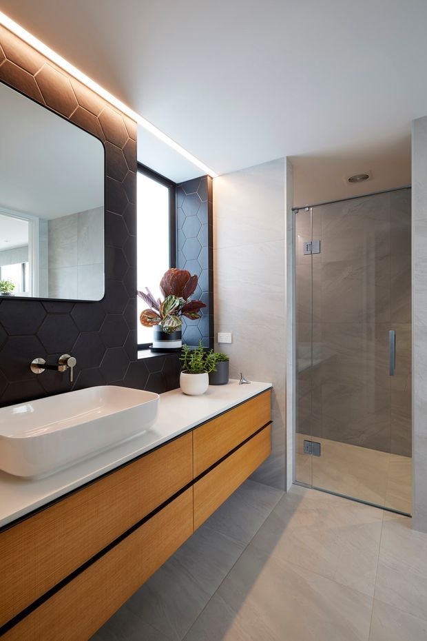 Contemporary bathroom featuring a floating timber vanity with a white vessel basin, large-format light grey floor and wall tiles, a black hexagonal feature-tile wall, frameless glass shower enclosure, black-framed window, and integrated warm LED lighting. A rounded-corner mirror and potted plants add softness to the refined, modern scheme.