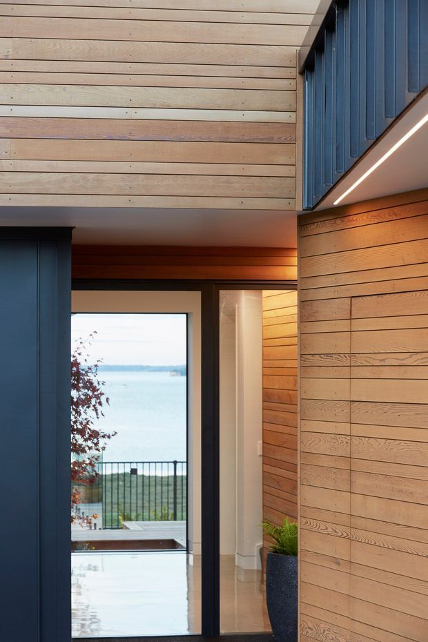 A refined residential interior passage with extensive horizontal timber wall and ceiling lining, dark blue vertical panelled joinery, recessed linear lighting, and large glazed openings leading to a balcony with a water view. The warm timber surfaces contrast with dark frames and cool blue cabinetry, creating a modern coastal atmosphere.