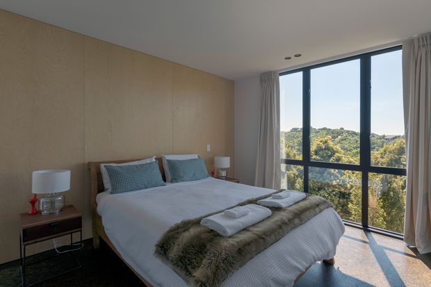 A bright, calm bedroom with a neatly made bed positioned against a full-height pale timber feature wall. The room includes bedside tables and lamps, layered white bedding, folded towels, and a textured faux-fur throw. Full-height black-framed glazing with curtains opens the view toward a leafy landscape, while recessed ceiling lights and a dark floor provide contrast.