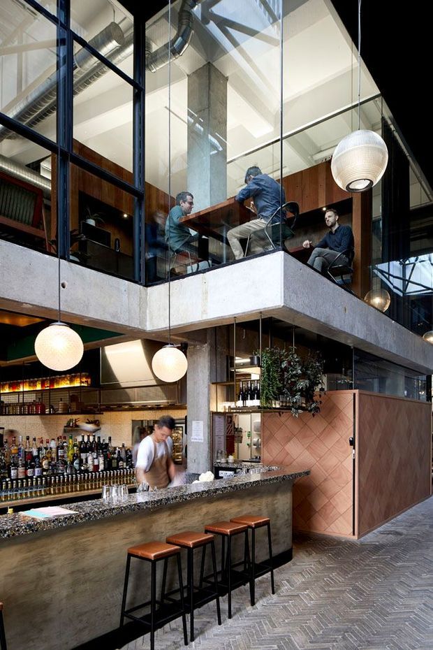 A vertically framed interior photograph of a multi-level restaurant or bar with a dramatic industrial-contemporary fit-out. The ground floor features a long concrete-faced bar with a speckled stone benchtop, liquor shelving, pendant globe lights, and leather-topped stools. Above, a mezzanine dining or lounge area is enclosed by black-framed glass balustrades and surrounded by exposed concrete columns, timber wall panels, and visible industrial ductwork. A herringbone-patterned floor and large glazed openings complete the layered, urban hospitality setting.