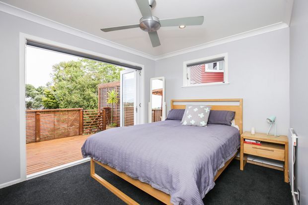 Bright contemporary bedroom with a timber platform bed, grey-purple bedding, dark carpet, pale grey walls, white trim, a ceiling fan, bedside table, and large sliding glass doors opening to a timber balcony or deck with privacy screening.