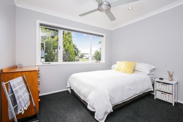 Bright, tidy bedroom with pale grey walls, white trim, a large window overlooking greenery, dark carpet, and a neatly made bed with white bedding and a yellow patterned accent cushion. A timber dresser, drying rack, ceiling fan, and small bedside shelving complete the room.