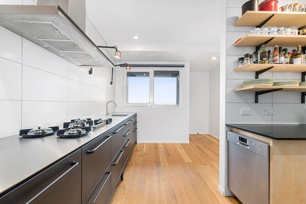 Bright, narrow contemporary kitchen with a long dark built-in cooktop run, stainless rangehood, white tiled walls, black benchtops, open timber shelving, integrated dishwasher, and warm timber flooring. A window brings in natural light at the far end.