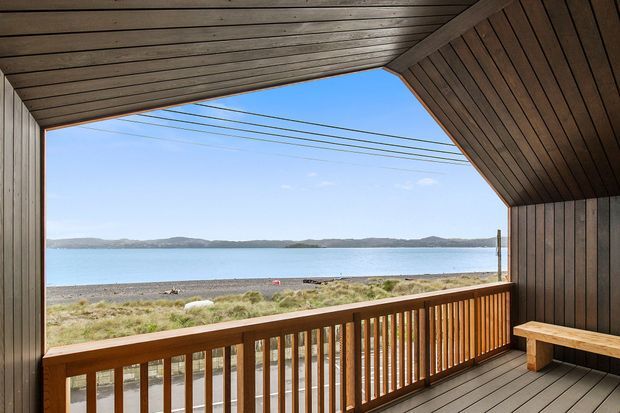 Covered timber-clad balcony or terrace with a dramatic angled timber ceiling, vertical timber wall panelling, timber decking, and a simple built-in bench. The opening frames a clear view over a beach, calm water, and distant low hills, with warm natural wood dominating the architectural finish.