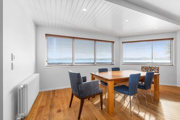 Bright dining room with a solid timber dining table and six upholstered chairs, warm timber flooring, large windows with timber Venetian blinds, white walls, and a white painted tongue-and-groove ceiling. A radiator and recessed ceiling lights are visible, creating a clean contemporary interior with warm natural accents.