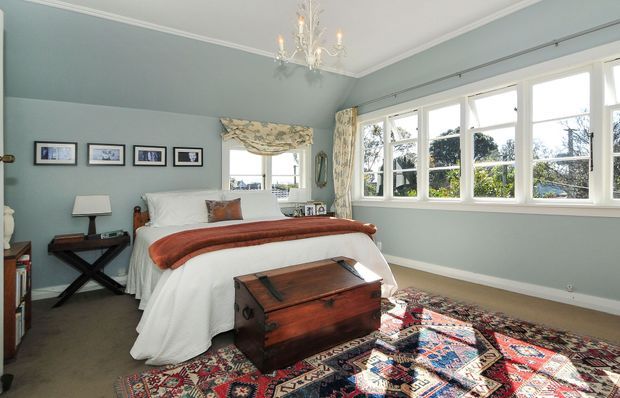 Bright traditional bedroom with pale blue-grey walls, white trim, a centered upholstered bed dressed in white linens and a rust-coloured throw, matching patterned curtains, framed artwork, a chandelier, a dark wood bedside table, a vintage wooden chest at the foot of the bed, and a richly patterned oriental rug. Large multi-pane windows provide abundant natural light and views of greenery.
